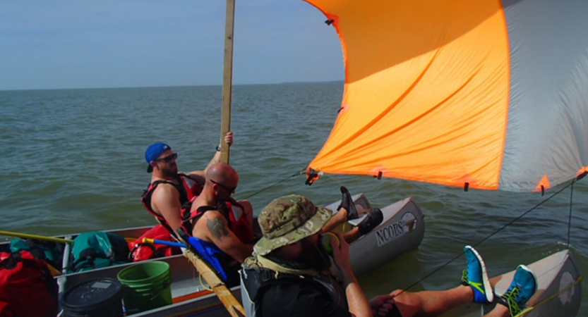 three people sit in canoes using a tarp as a makeshift sail